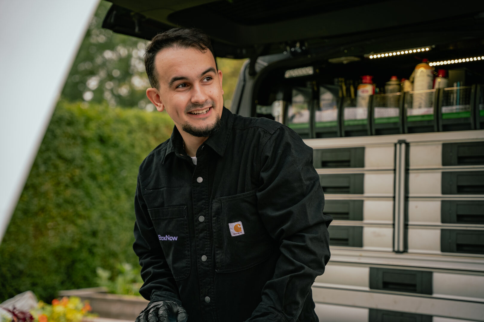 En mand i sort skjorte smiler, mens han arbejder på en bil. Bagved er en varevogn fyldt med værktøj og materialer, omgivet af grønne buske. Tekst: "BoxNow".