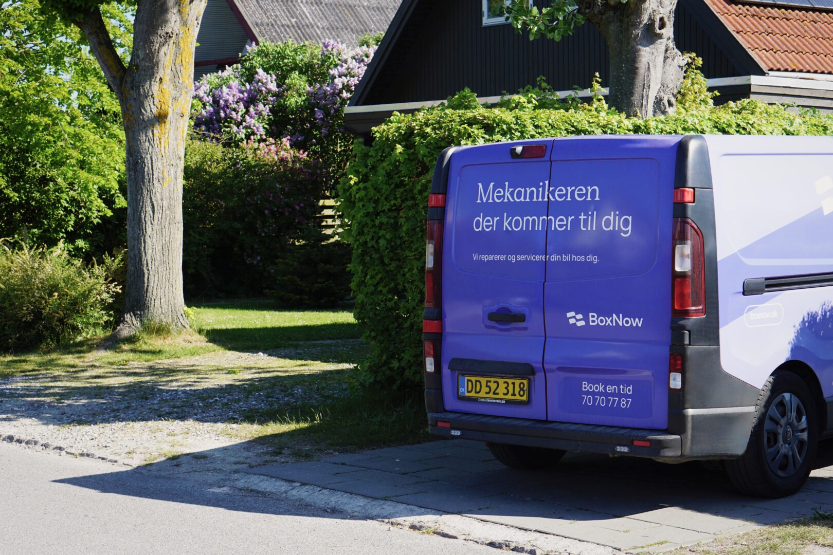 Bagenden af BoxNow servicebil med budskabet “Mekanikeren der kommer til dig” parkeret foran dansk parcelhus