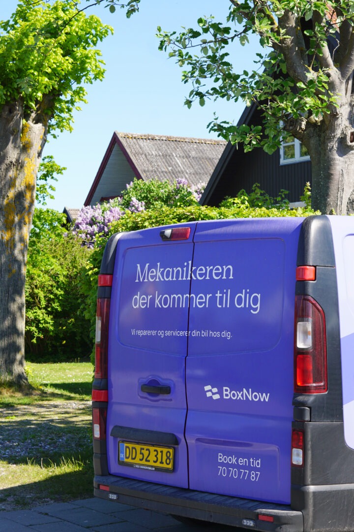 Bagenden af BoxNow servicebil med teksten “Mekanikeren der kommer til dig” parkeret foran parcelhus klar til mobil bilservice
