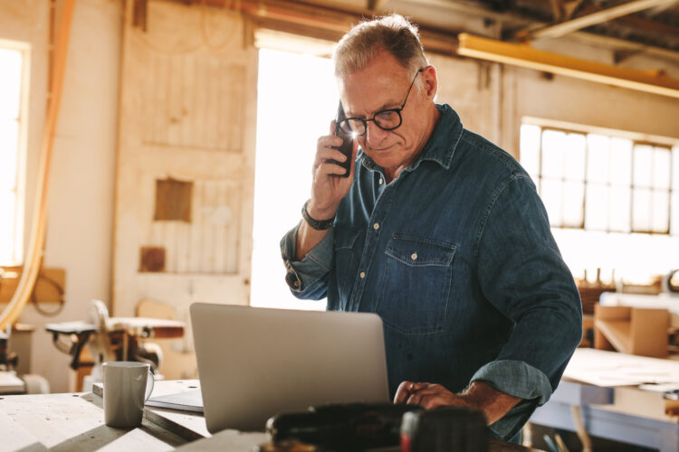 En mand i denimskjorte taler i telefon og arbejder på en bærbar computer i et lyst værksted fyldt med værktøj og træudstyr. En kaffekop står på bordet.