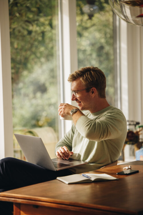 En person sidder ved et bord med en bærbar computer, smiler og drikker fra en kop. Rummet er fyldt med naturligt lys fra store vinduer, og der er en notesbog og høretelefoner på bordet.