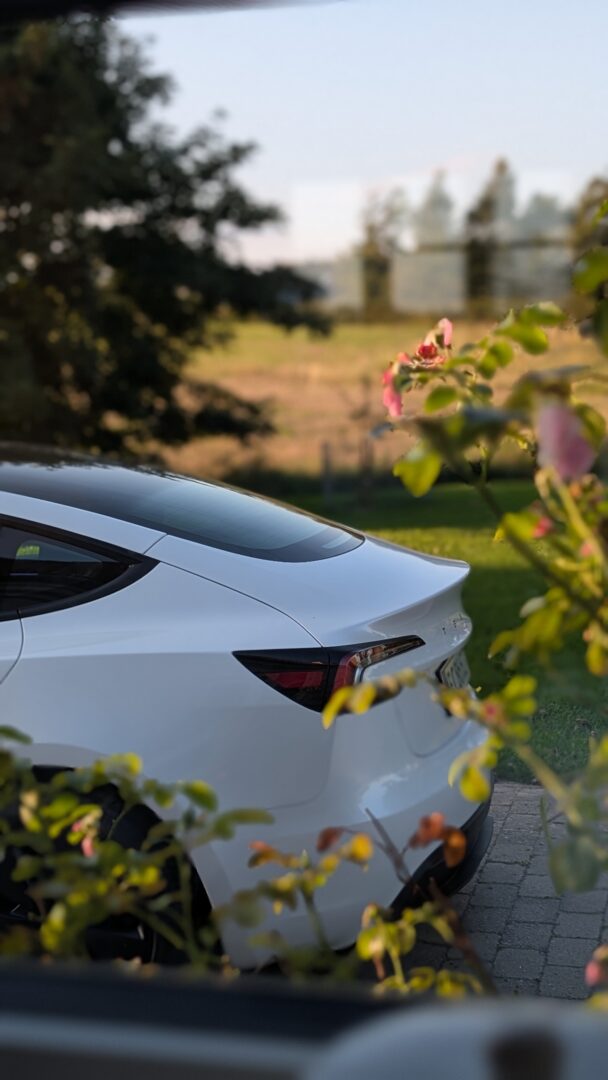 Hvid Tesla parkeret i en indkørsel omgivet af grønne planter og blomster. Klar til service direkte hjemme hos dig.
